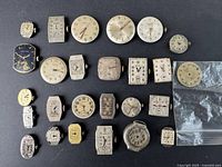 Photo showing 23 vintage watch movements with dials in various shapes and colors laid out on a black surface