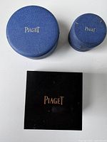Three vintage Piaget branded watch and jewelry cases of different size and shape, two blue leatherette round cases and one square black case.