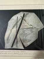 Close-up image showing the mixed media artwork with abstract geometric shapes in grey and black on paper, framed and with handwritten text below the image area.