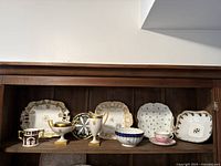 Photo showing the full set of antique china pieces on a wooden shelf, including plates, cups, a creamer, and sugar bowl with gold accents.