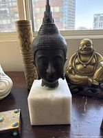 Black Buddha head sculpture on marble base with brass laughing Buddha and carved container in background
