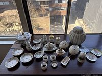 Wide view of entire set on table by window showing various lidded containers, salt shaker, plates and small cups in floral and fruit-themed painted ceramic and porcelain.