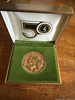 Medallion seated in its hinged presentation box, lid open showing printed “50” graphic on satin-lined interior and green velvet base.