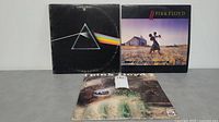 Three Pink Floyd LP records shown together, including The Dark Side of the Moon, another with couple dancing on a farm cover, and another with surreal face and green colors on cover.
