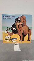 Front cover of 'Mystery To Me' album by Fleetwood Mac showing cartoon dog on beach with cake and album title.