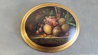 Full view of the oval wall hanging painting depicting fruit on a table, showing gold frame and overall condition.
