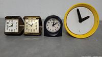 Four alarm clocks on gray surface: two Westclox travel alarm clocks with cases open, one small black Bulova alarm clock, one round yellow clock with minimalist black hands.