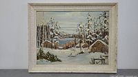 Front view of framed winter scene painting with snow-covered cabins, trees, mountains, lake, and cloudy sky.