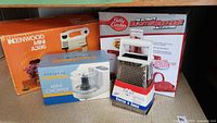 Lot showing Betty Crocker Blaster Blender box, Kenwood Mini A386 box, Everyday Essentials Mini Chopper box, and a hand grater/slicer set on a wooden surface.