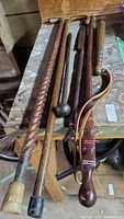 Photo showing six wooden canes and sticks laid out on a workbench with visible wear and patina.