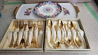 Full view of 6 gold forks and 6 gold spoons displayed in lined boxes with a rectangular China tray and small round China plate behind them.