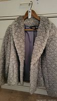 Front view of light grey faux fur jacket on hanger showing scalloped fur pattern and folded collar.