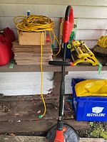 Red and black Grass Hog electric weed wacker leaning against a wall with other gardening supplies including stacked yellow garden bags, red fuel containers, and yellow garden accessory parts.