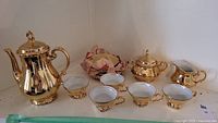 Full view of coffee pot, 5 coffee cups with saucers, cream jug, and sugar bowl with lids, all gold coloured and reflective