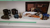 Two black mugs with gold classical Greek designs, religious icon plaque and a blue animal figure, large beads in tray, and hammered copper plate in background