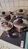 Four Mid-Century Modern styled metal saucepans with lids on a stove, featuring geometric orange and black patterns on the sides.