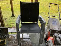 Photo showing one black outdoor chair with metal frame and mesh seat and back, placed on outdoor wooden deck with lawn mower in background.