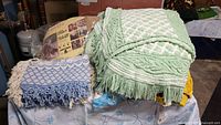 Stack of vintage blankets including a green chenille with fringe and blue and white woven blanket with tassels.