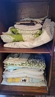Stack of folded vintage linens on wooden shelves showing mix of lace, cotton and patterned fabrics in green, white and cream tones