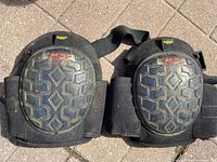 Pair of black knee pads with gel cushioning and patterned outer shell, elastic Velcro straps, placed on a tiled floor