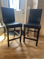 Two wood and black pleather tall bar stools photographed side by side near a door with a wood floor.