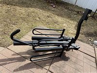 Full image of the folded black vehicle-mount bike rack on a backyard patio with grass background showing support arms and hitch connection.
