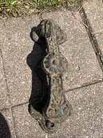 Top angle photo showing floral and ornate pattern on black cast iron door knocker laid on concrete surface with some grass nearby.