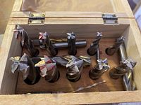 Top-down view of fifteen large metal drill bits of varying shapes, stored upright in slots inside a wooden box with metal hinges and latch.
