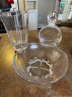 Clear glass bowl with heart shape design visible at the base, placed on a countertop next to a round decanter and a tall glass vase.