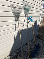 Two metal fan rakes with wooden handles leaning against a wall alongside the Yardworks snow shovel with blue handle.