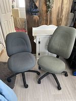 Photo showing two fabric-upholstered rolling desk chairs, one green with high back and one gray with lower back, both with caster wheels and adjustment features.