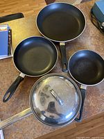 Set of three non-stick frying pans and one pot with lid on table, showing black cooking surfaces and plastic handles, and reflective metal pot lid.