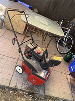 Snow blower shown from angled side highlighting handle and engine controls with a glass table and bike in background