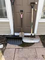 Full view of three shovels including large plastic snow shovel, metal garden shovel, and smaller metal snow shovel, all used with wood handles.