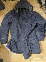 Full view of navy blue men's winter coat with hood laid flat on tile floor showing front details including pockets and sleeve logo.