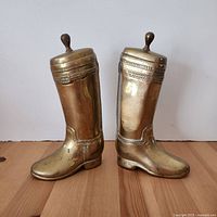 Front-side view of two solid brass boot bookends, both left feet with knob tops, showing detailed stitch and buckle designs.