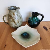 Three ceramic pottery pieces: speckled jug with repaired handle on left, dark green jug on the right, small cream square trinket plate with glaze spot in front center on wooden surface.