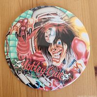 Front view of large circular Mötley Crüe button showing detailed cartoon art and band logo on wooden surface.