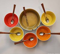 Set of six ceramic condiment bowls; five smaller in red, yellow, and orange with wooden-handled spoons, one larger olive green dish with fork.