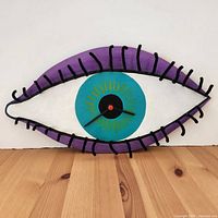 Full frontal view of the folk art eye clock showing the entire eye shape, purple eyelid frame with black yarn eyelashes, and the centrally located clock face.