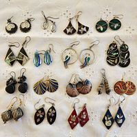 Photo showing full lot of 17 pairs of vintage earrings arranged on white embroidered fabric backdrop, various styles including Mexican silver and abalone shell.