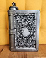 Front side of pewter book flask with ornate shield and floral scroll design