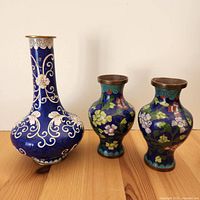 Front view of three cloisonné vases including large blue vase with white floral scroll design and two smaller matching bud vases with colorful floral patterns.
