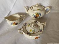 Photo showing the three-piece porcelain set of teapot, creamer, and sugar pot with floral patterns and gold trim on cream background, arranged on white surface.