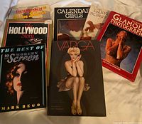 Seven coffee table books stacked and arranged on a white surface, titles visible including 'Varga', 'Hollywood Story', 'Calendar Girls', 'The Best of 10 Modern Screen', 'Encyclopedia of Film Stars', 'Erotic Images', and 'Glamour Photography Techniques'.