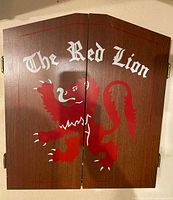 Closed view of wooden dart cabinet with painted red lion and 'The Red Lion' text on the outside of two hinged doors.
