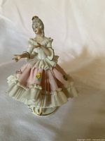 Front angled view of a Dresden porcelain figurine showing intricate lace details on the dress and small floral decorations, painted in pink, white, and gold.