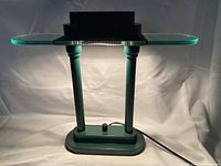 Front view of green glass and metal desk lamp illuminated showing green glass top and metal supports with base and light housing visible beneath glass.