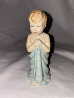 Front view of child figurine with hands clasped and wrapped in light blue towel, showing facial detail and patterning on towel.