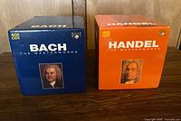Two boxed CD sets side by side, one blue Bach set and one orange Handel set showing front portraits and titles.